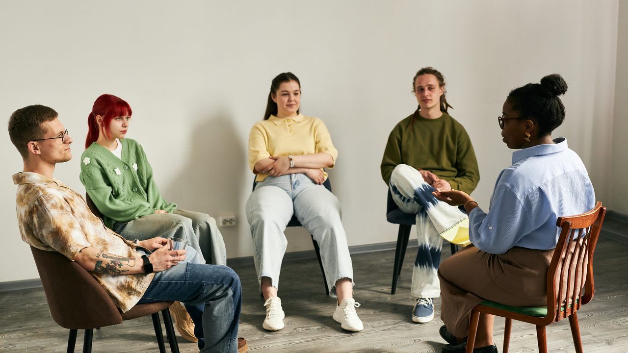 A small support group of adults seated in a sunlit indoor space, mid-conversation.