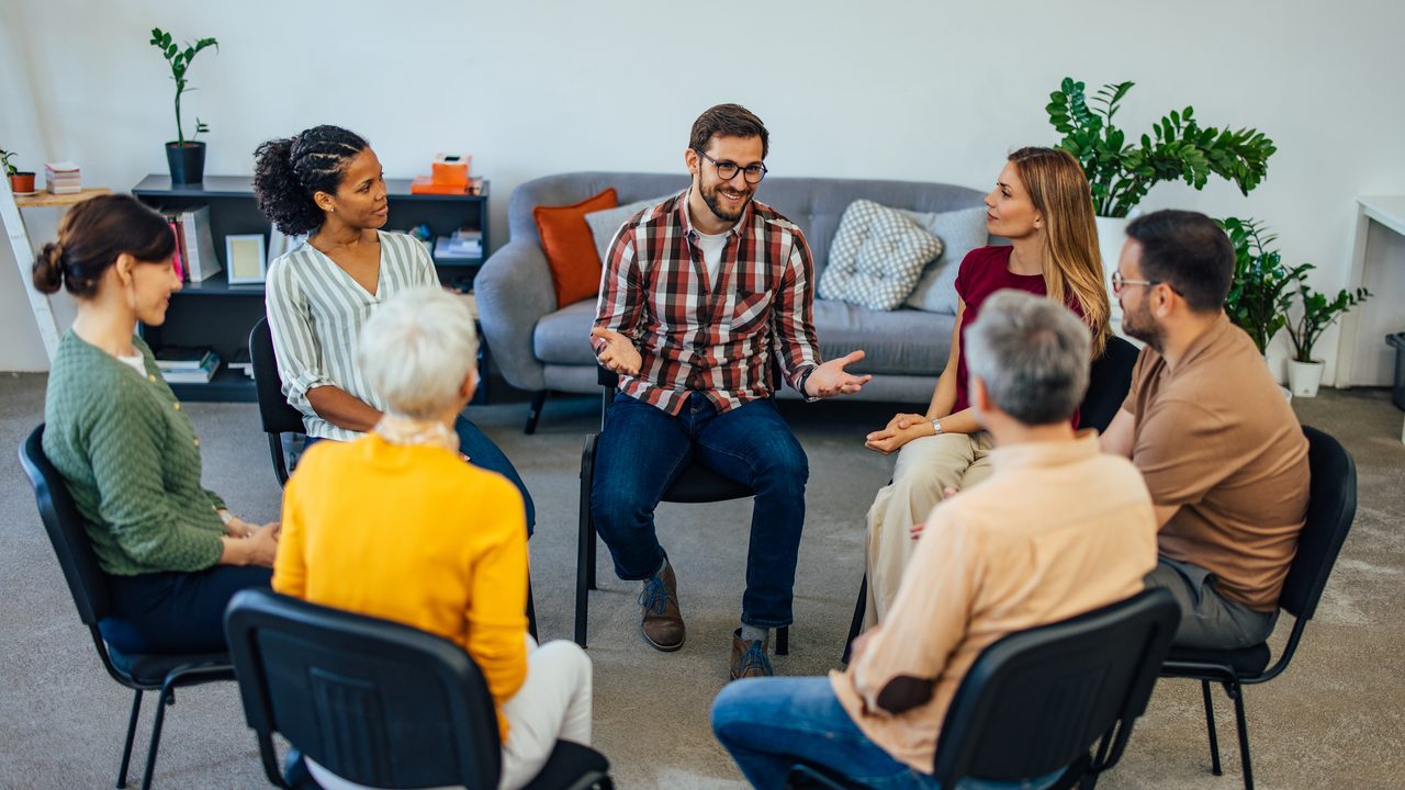 A small support group sitting together in a bright living room, mid-conversation, with sunlight on the floor.